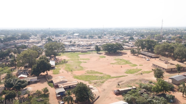 An aerial view of a vast, open area with patches of grass and dry soil. The landscape includes several buildings scattered around the foreground and more densely packed structures in the background, suggesting a community or town. Trees are interspersed throughout the area, providing greenery. A few people can be seen walking or standing in the open field.