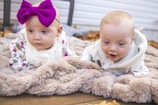 Two twin baby girls playing with colorful toys on a soft rug.
