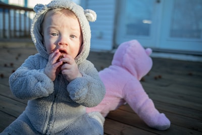 A baby wearing a grey, bear-themed onesie with a hood sits on a wooden deck, holding something close to its mouth. In the background, another baby in a pink, similarly themed onesie is crawling away from the camera. The environment appears to be an outdoor porch with white railings and a blue door in the background.