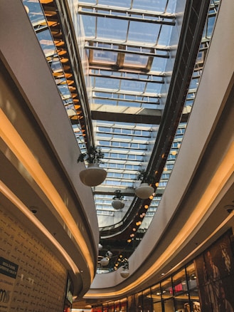 A modern indoor shopping mall with large glass windows in the ceiling allowing natural light to flood the space. Curved architectural lines and suspended round planters create a sleek and contemporary atmosphere. Electric lighting illuminates the mall with warm tones, complementing the natural light.