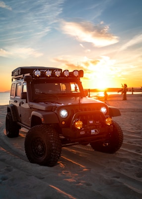 A rugged Jeep with large off-road tires is parked on a sandy beach. It is equipped with roof lights and bull bars, and the sunset in the background casts a warm glow over the scene. Palm trees and the ocean are visible in the distance, contributing to a serene atmosphere.