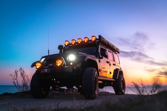 A rugged off-road vehicle with large tires and roof-mounted lights is parked on a dirt path, set against a dramatic sunset. The sky displays a gradient of pink, orange, and blue hues, adding a sense of adventure to the scene.