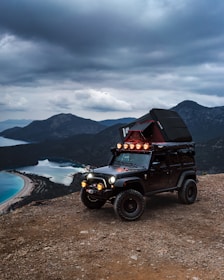 A rugged off-road vehicle equipped with a rooftop tent parked on a rocky terrain. The vehicle is placed against a backdrop of dramatic, cloudy skies and overlooking a serene body of water with mountains in the distance. The scene suggests a sense of adventure, with the vehicle's lights turned on, enhancing the atmosphere.