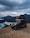 A rugged off-road vehicle equipped with a rooftop tent parked on a rocky terrain. The vehicle is placed against a backdrop of dramatic, cloudy skies and overlooking a serene body of water with mountains in the distance. The scene suggests a sense of adventure, with the vehicle's lights turned on, enhancing the atmosphere.