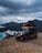 A rugged off-road vehicle equipped with a rooftop tent parked on a rocky terrain. The vehicle is placed against a backdrop of dramatic, cloudy skies and overlooking a serene body of water with mountains in the distance. The scene suggests a sense of adventure, with the vehicle's lights turned on, enhancing the atmosphere.