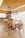 A modern dining room features a long wooden table surrounded by stylish wooden chairs. Above the table hang two contemporary rectangular pendant lights with exposed bulbs. The room is decorated with neutral tones and has a minimalist design. In the background, there is a kitchen area with wooden cabinets, a built-in oven, and various kitchenware items.