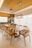 A modern dining room features a long wooden table surrounded by stylish wooden chairs. Above the table hang two contemporary rectangular pendant lights with exposed bulbs. The room is decorated with neutral tones and has a minimalist design. In the background, there is a kitchen area with wooden cabinets, a built-in oven, and various kitchenware items.