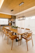 A modern dining room features a long wooden table surrounded by stylish wooden chairs. Above the table hang two contemporary rectangular pendant lights with exposed bulbs. The room is decorated with neutral tones and has a minimalist design. In the background, there is a kitchen area with wooden cabinets, a built-in oven, and various kitchenware items.