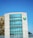 A modern building features a large facade of glass windows and a distinctive circular green logo on the wall. The structure is partially adjacent to another section with a checkered pattern of neutral tones. The sky is clear and blue, contributing to a clean and contemporary aesthetic.