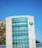 A modern building features a large facade of glass windows and a distinctive circular green logo on the wall. The structure is partially adjacent to another section with a checkered pattern of neutral tones. The sky is clear and blue, contributing to a clean and contemporary aesthetic.