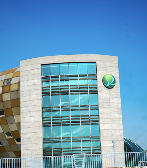 A modern building features a large facade of glass windows and a distinctive circular green logo on the wall. The structure is partially adjacent to another section with a checkered pattern of neutral tones. The sky is clear and blue, contributing to a clean and contemporary aesthetic.