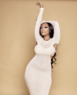 A person with long, wavy hair poses gracefully against a light brown backdrop, wearing a form-fitting, sheer white dress. Their arms are elegantly raised above their head, with delicate green nail polish visible. The expression is calm and composed, enhanced by subtle, polished makeup.