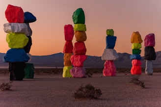 Tall stacks of brightly colored boulders are artfully arranged in a desert landscape against a backdrop of mountains and a sunset sky. Each rock is painted in vibrant colors, creating a striking contrast with the natural surroundings.