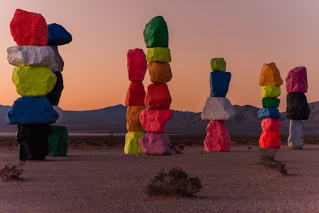 Tall stacks of brightly colored boulders are artfully arranged in a desert landscape against a backdrop of mountains and a sunset sky. Each rock is painted in vibrant colors, creating a striking contrast with the natural surroundings.