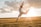 A person is captured mid-leap, executing a graceful jump against the backdrop of a glowing sunset. They are wearing a light, flowy outfit, and their posture suggests freedom and joy. The sky is partly cloudy, and the landscape is an expansive field, bathed in golden light.