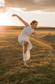 A person in a white dress is performing a graceful dance move on a grassy field during sunset. The sunlight casts a warm glow and highlights the dancer's movements, creating a serene and artistic scene.