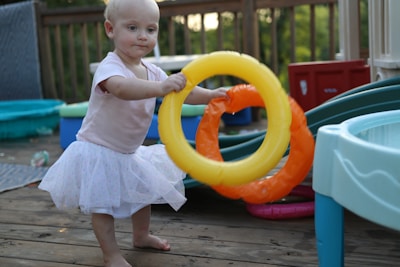 A young child wearing a light pink shirt and a white tutu skirt is playing with inflatable rings on a wooden deck. The child looks focused while holding a yellow and an orange inflatable ring. Surrounding the child are various play items including a slide, small pools, and a blue table. The setting is outdoors, possibly in a backyard with a wooden fence and green foliage in the background.