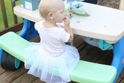 A young child with short blond hair is seated on a green bench, wearing a white shirt and a light, translucent tutu with polka dots. The child is eating a small snack, and a plastic table in front holds a partially visible food container.