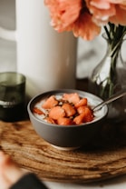 A bowl filled with yogurt topped with sliced strawberries and a sprinkle of nuts or seeds sits on a wooden surface. A spoon is placed inside the bowl, ready for eating. In the background, a blurred vase holds pink flowers, and there is a small candle.