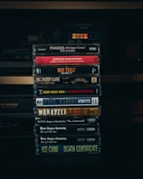 A collection of cassette tapes is stacked in front of an old stereo system. Each cassette features classic hip-hop albums from the 1990s, including artists like Fugees, Arrested Development, Mad Skillz, Big Daddy Kane, Snoop Doggy Dogg, Dr. Dre, Makaveli, Bone Thugs-n-Harmony, and Ice Cube.