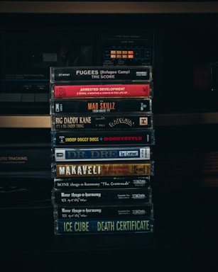 A collection of cassette tapes is stacked in front of an old stereo system. Each cassette features classic hip-hop albums from the 1990s, including artists like Fugees, Arrested Development, Mad Skillz, Big Daddy Kane, Snoop Doggy Dogg, Dr. Dre, Makaveli, Bone Thugs-n-Harmony, and Ice Cube.