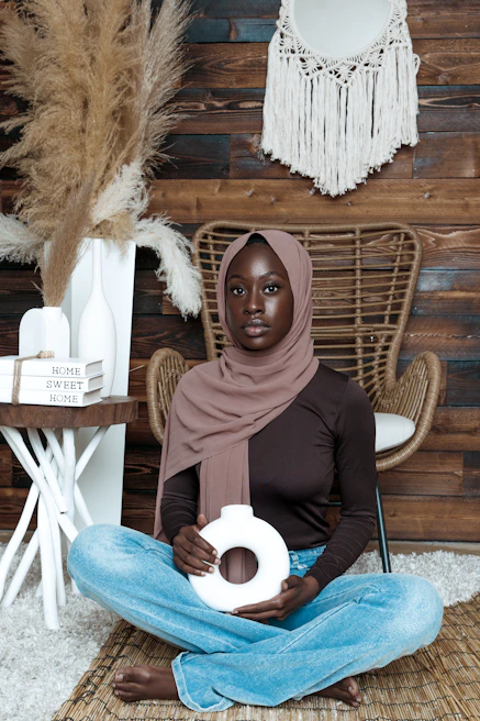 A person wearing a brown hijab and dark shirt is sitting cross-legged on a textured rug. They hold a white circular object. The background features a wicker chair, a wooden wall, a macramé wall hanging, and pampas grass placed in a white vase. Nearby, there is a small table with stacked books labeled 'Home Sweet Home'.