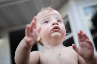 A young child with a curious expression, reaching forward with both hands. The child's face is partially smudged with dirt, creating a playful and adventurous impression. The background suggests an outdoor setting, possibly a backyard or garden.