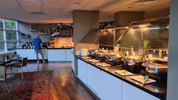 A buffet area with several covered dishes arranged on a counter. A person in a blue shirt and shorts is preparing a plate at the far end, near a display of baked goods. The room has wooden floors and modern decor, with a patterned rug and green plants visible on the counters.