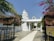 A white Hindu temple structure with ornate carvings is visible, surrounded by lush greenery and colorful flowers. The open gates to the temple are adorned with decorations, and a pathway leads up to the main building. Palm trees and flowering plants add to the serene atmosphere.
