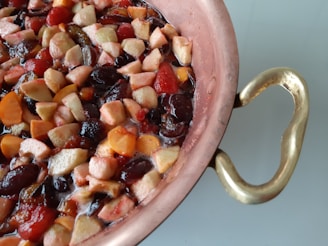 A copper pot filled with a colorful mixture of chopped fruits, including apples, grapes, and berries, creating a vibrant fruit medley. The pot has a detailed brass handle, adding a touch of elegance.