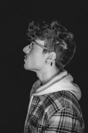 A black and white profile portrait of a person wearing glasses. The individual has tousled hair, a plaid jacket over a hoodie, and an earring with a star-shaped design. The background is dark, creating a strong contrast with the subject.