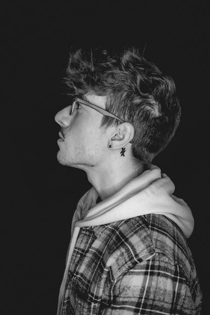 A black and white profile portrait of a person wearing glasses. The individual has tousled hair, a plaid jacket over a hoodie, and an earring with a star-shaped design. The background is dark, creating a strong contrast with the subject.