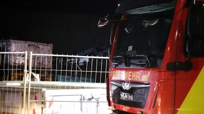 A fire truck parked near a construction site or building, visible through a metal fence. The truck is red and yellow with signage indicating it's a fire service vehicle. The surrounding area is dimly lit, highlighting part of the truck and some construction materials.