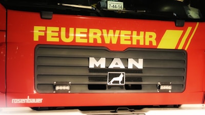 Front view of a red fire truck with bold yellow lettering 'FEUERWEHR' across the top of the vehicle. Below the lettering, the brand name 'MAN' is prominently displayed in silver on a black grille. The lower part of the grille includes a logo of a lion. The truck has additional branding 'rosenbauer' in white at the bottom left of the image.