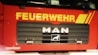 Front view of a red fire truck with bold yellow lettering 'FEUERWEHR' across the top of the vehicle. Below the lettering, the brand name 'MAN' is prominently displayed in silver on a black grille. The lower part of the grille includes a logo of a lion. The truck has additional branding 'rosenbauer' in white at the bottom left of the image.