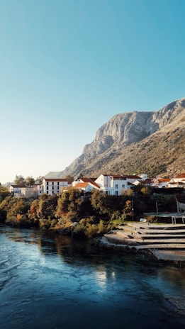 A serene riverside village is nestled at the foot of a towering mountain. The village features charming houses with red roofs surrounded by lush green trees. The river in the foreground reflects the clear blue sky and the light gently illuminates the landscape, providing a peaceful and picturesque scene.