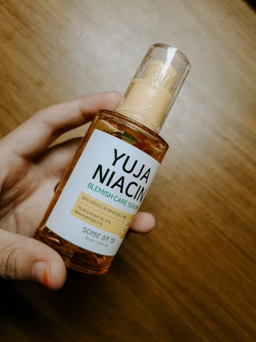 A hand is holding a bottle of YUJA NIACIN Blemish Care Serum, which is labeled with details about its ingredients such as Yuja Extract and Niacinamide. The bottle has a translucent design with a pump dispenser, and it is placed against a wooden background.
