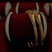 Two intricately designed gold bangles are displayed against a luxurious red velvet background, highlighting the jewelry's elegance and craftsmanship.
