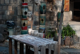 A rustic outdoor scene features a wooden table with several white ceramic cups placed on it. Behind the table, there are multiple large, colorful, cylindrical pottery pieces stacked against a stone wall. The area is partly shaded with green plants in decorative pots placed along the wall.
