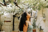 A woman stands in a garden with lush green vines overhead. Ambient lighting and natural wooden beams create a calm, outdoor setting. The walls are made of stone, and various potted plants add to the tranquil environment.