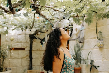 A woman stands in a garden with lush green vines overhead. Ambient lighting and natural wooden beams create a calm, outdoor setting. The walls are made of stone, and various potted plants add to the tranquil environment.