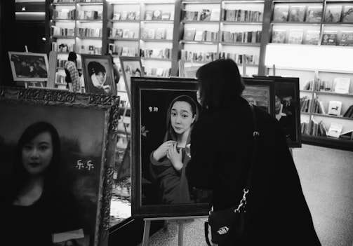 Black and white photograph of a gallery or bookstore with several framed portraits on display. A person is closely examining one of the portraits, which depicts a woman with her hands clasped. In the background, bookshelves and more artworks are visible.