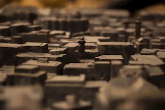 A close-up view of a detailed miniature model of a city with various buildings and structures, mostly composed of dark tones. The depth of field creates a blurred background effect, emphasizing the textured surfaces of the miniature buildings.