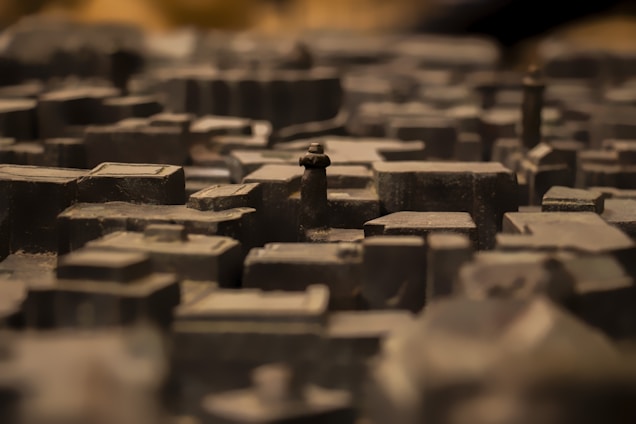 A close-up view of a detailed miniature model of a city with various buildings and structures, mostly composed of dark tones. The depth of field creates a blurred background effect, emphasizing the textured surfaces of the miniature buildings.