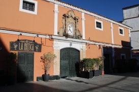 A historic building with an orange fa&ccedil;ade features a large, ornate central door and decorative stonework. The establishment, named 'La Botica', has a sign with traditional design. Potted plants are placed on either side of the entrance, and the building is set against a clear blue sky.