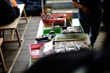 A table is covered with various food items packaged in plastic containers, trays, and baskets. The table is set in a casual setting with wooden benches nearby. Light snacks and wrapped food items are organized across different colored containers, and there is a person partially visible on the right side.