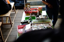 A table is covered with various food items packaged in plastic containers, trays, and baskets. The table is set in a casual setting with wooden benches nearby. Light snacks and wrapped food items are organized across different colored containers, and there is a person partially visible on the right side.