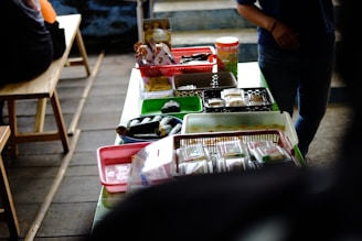 A table is covered with various food items packaged in plastic containers, trays, and baskets. The table is set in a casual setting with wooden benches nearby. Light snacks and wrapped food items are organized across different colored containers, and there is a person partially visible on the right side.