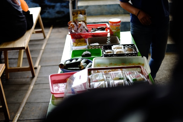 A table is covered with various food items packaged in plastic containers, trays, and baskets. The table is set in a casual setting with wooden benches nearby. Light snacks and wrapped food items are organized across different colored containers, and there is a person partially visible on the right side.