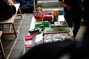 A table is covered with various food items packaged in plastic containers, trays, and baskets. The table is set in a casual setting with wooden benches nearby. Light snacks and wrapped food items are organized across different colored containers, and there is a person partially visible on the right side.